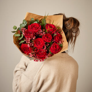 Person holding a bouquet of red roses wrapped in brown paper against a neutral background